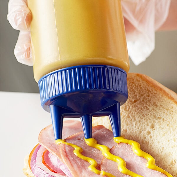 A hand using a Vollrath Tri Tip squeeze bottle with a blue cap to put mustard on a sandwich.