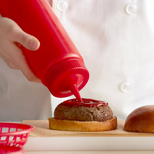 A hand using a Vollrath red wide mouth squeeze bottle to pour ketchup onto a hamburger.
