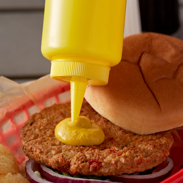 A burger with mustard being poured from a Vollrath yellow squeeze bottle.
