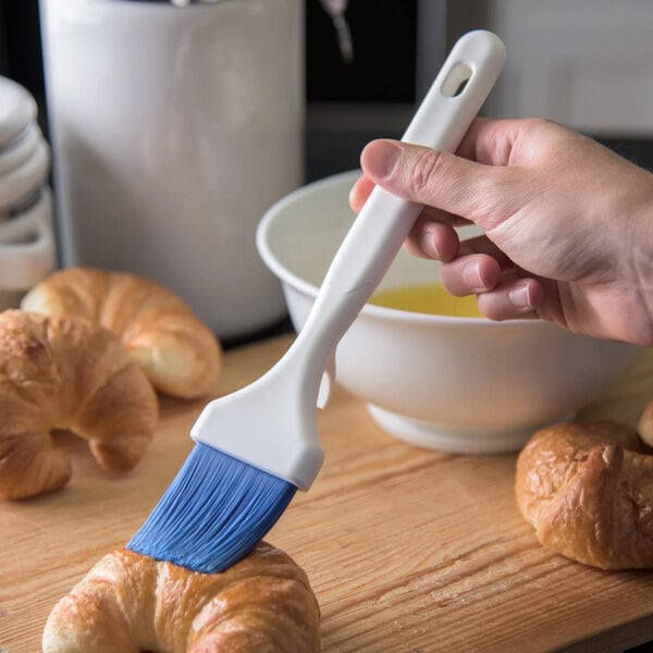 A hand using a blue Carlisle Sparta Spectrum basting brush to apply liquid to a croissant.