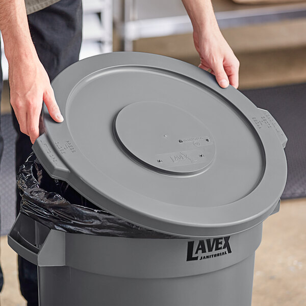 A person opening a Lavex gray round commercial trash can lid.