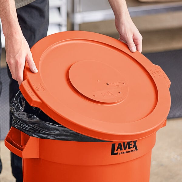 A person holding a Lavex orange plastic lid over a garbage can.