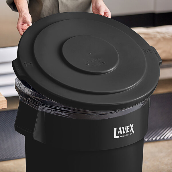 A person holding a Lavex black round commercial trash can lid over a black trash can.