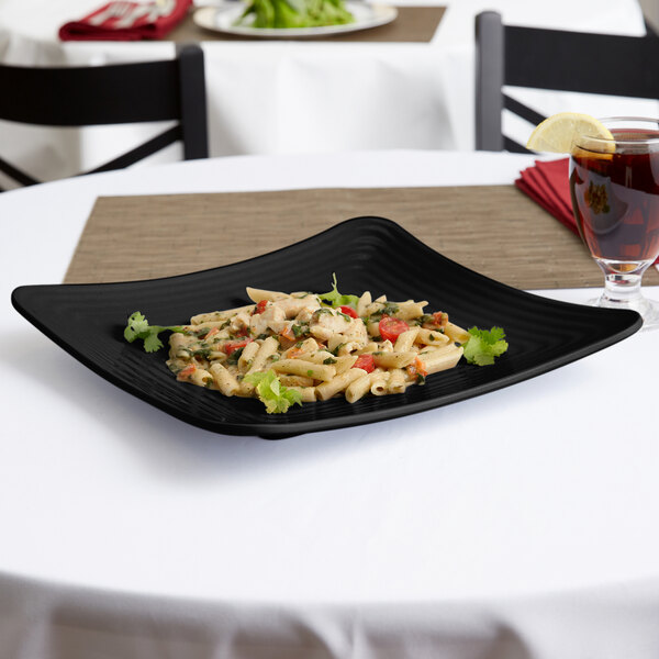 A black square Milano melamine plate with pasta and a drink on a table.