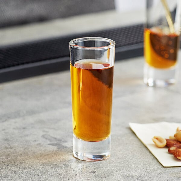 An Acopa shot glass filled with brown liquid and nuts on a table.