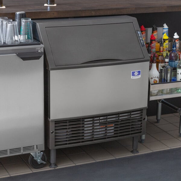 A Manitowoc stainless steel undercounter ice machine with a white lid on a counter.
