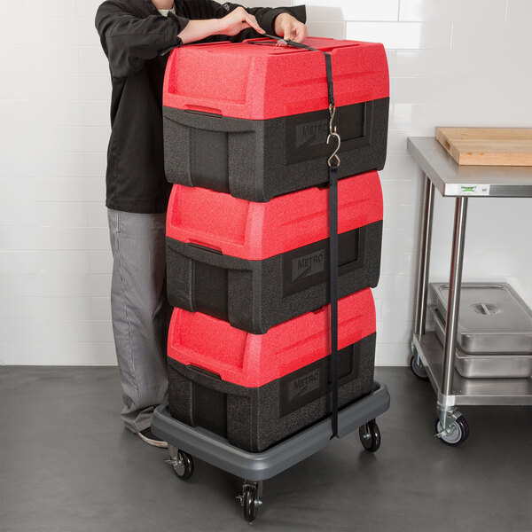 A man pushing a stack of red and black Metro Mightylite BigBoy food pan carriers on a dolly.