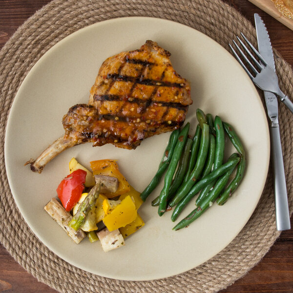 A Tuxton Zion Matte Beige china plate with grilled pork chops, green beans, and grilled vegetables on a table.