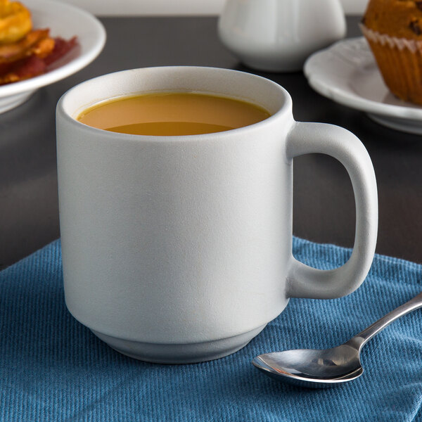 A white Tuxton china mug filled with coffee sitting on a blue cloth next to a muffin.