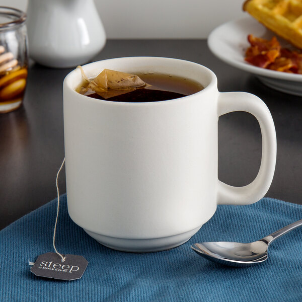 A Tuxton matte white china mug filled with tea with a tea bag in it on a blue surface with a spoon.