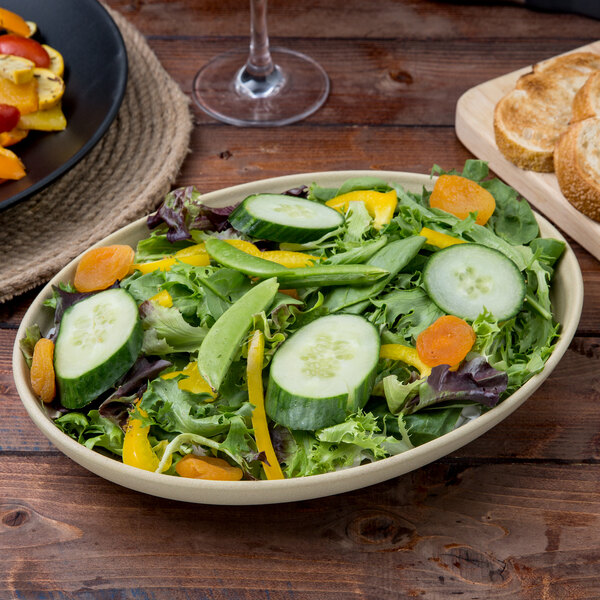 A Tuxton TuxTrendz matte beige bowl filled with salad with cucumbers and peppers next to a plate of bread.