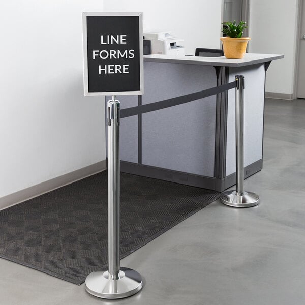 A Lancaster Table & Seating crowd control stanchion with a sign that says "line forms here" over a line of people.