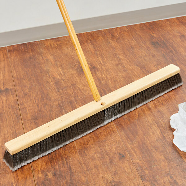 A Carlisle push broom head with gray flagged bristles on a wood floor.