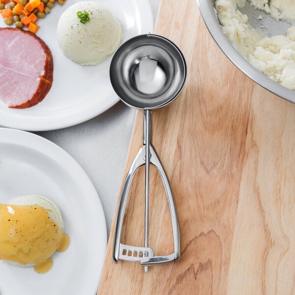 A silver Vollrath ice cream scoop with a squeeze handle on a table with a bowl of mashed potatoes.