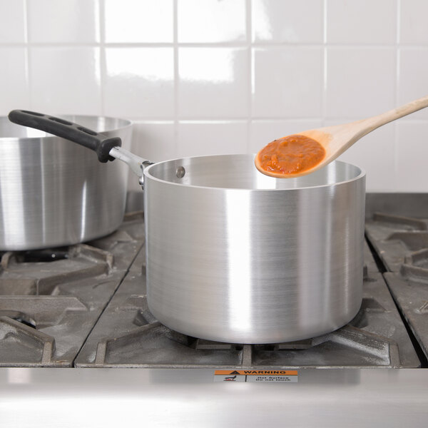 A wooden spoon stirring red sauce in a Vollrath Wear-Ever aluminum sauce pan on a stove.