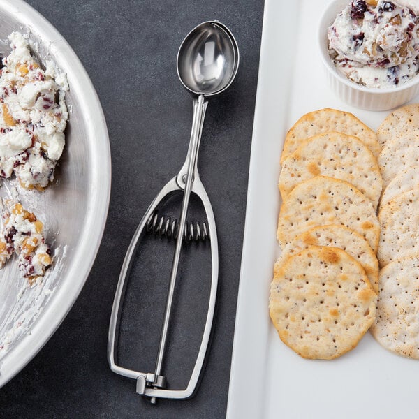 A plate of crackers with a bowl of cream cheese served with a Vollrath stainless steel squeeze handle disher.