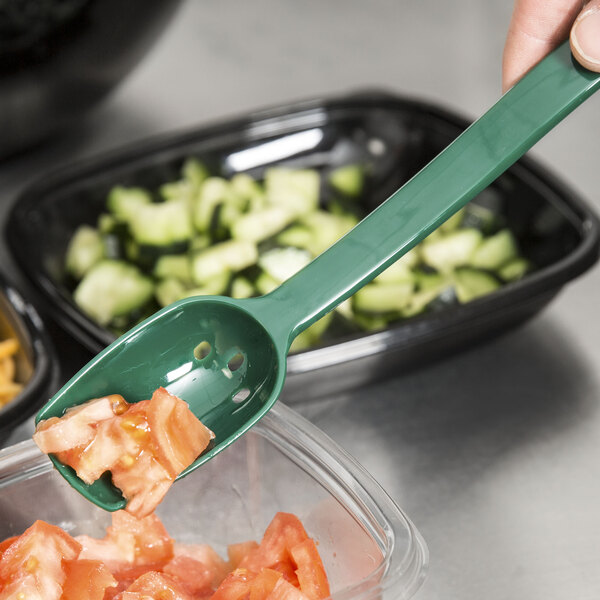 A hand holding a green Thunder Group perforated salad bar spoon filled with food over a bowl of tomatoes.