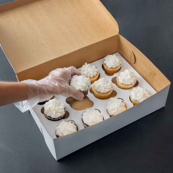 A person wearing plastic gloves reaching out to take a white 14" x 10" cupcake box with a clear window and 12 cupcakes inside.