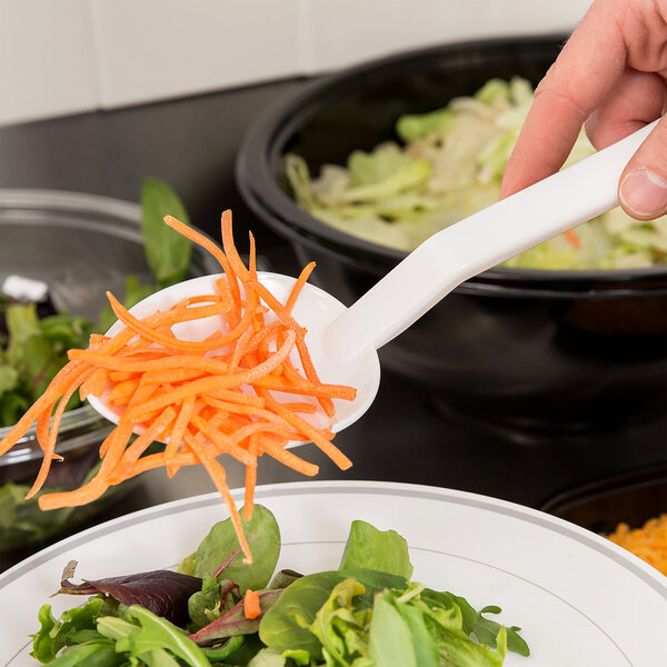 A person holding a Thunder Group white polycarbonate salad bar spoon filled with carrots over a plate of lettuce and carrots.