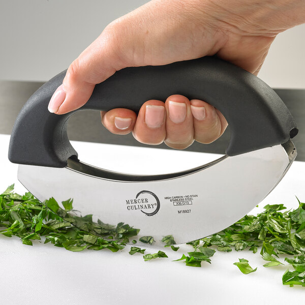 A hand holding a Mercer Culinary double blade mezzaluna knife with green herbs on it.