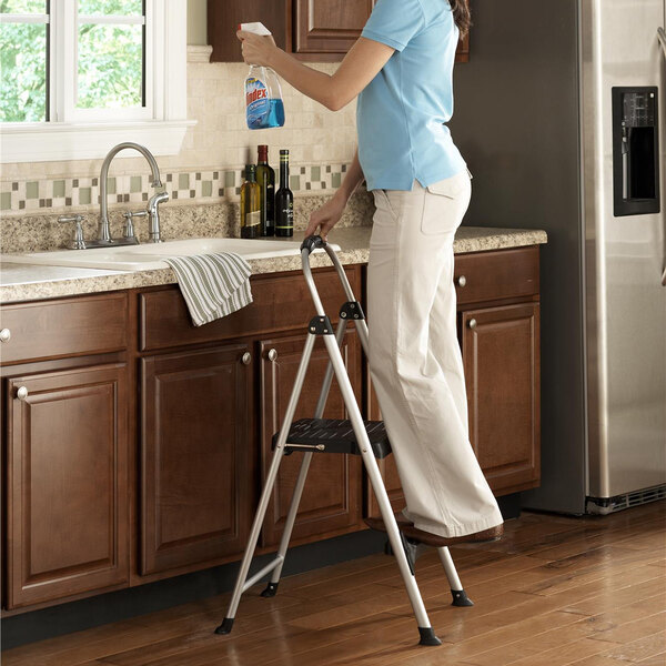 A woman standing on a Cosco two-step folding step stool in a kitchen.
