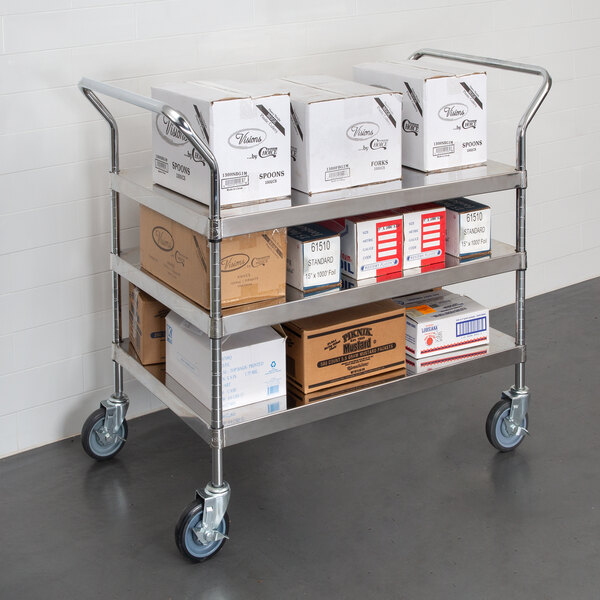 A Regency stainless steel three shelf utility cart with white boxes with black text on them.