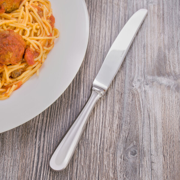 A silver Oneida Baguette dinner knife on a table with a plate of spaghetti and meatballs.