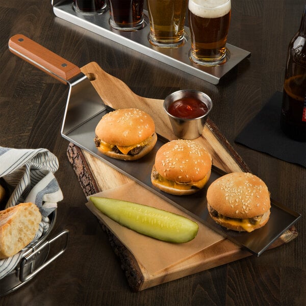 A Tablecraft stainless steel spatula serving a cheeseburger with a tray of cheeseburgers and a pickle on a wooden board.
