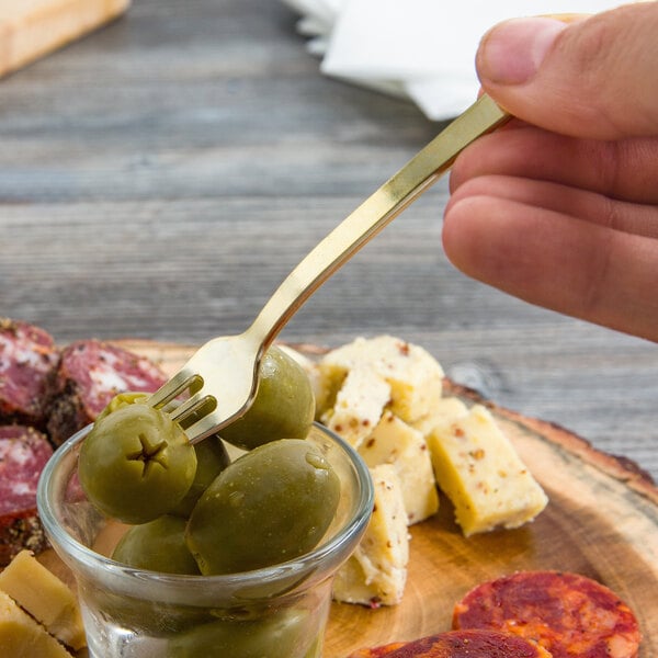 A hand holding a Fineline gold plastic tasting fork over a plate of olives and cheese.
