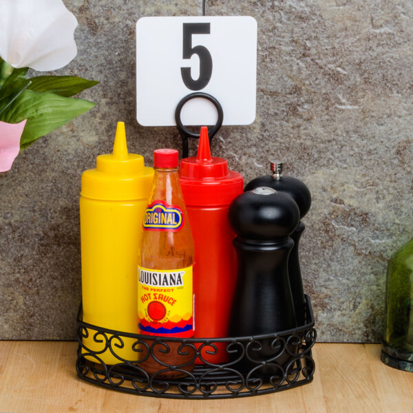 A Tablecraft Mediterranean black half condiment caddy holding a bottle of yellow mustard and a bottle of hot sauce on a table.