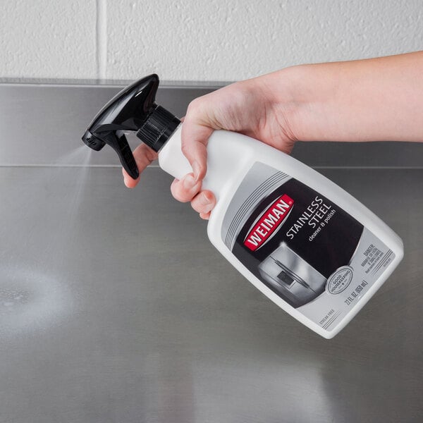 A hand holding a Weiman stainless steel cleaner and polish spray bottle over a stainless steel counter.