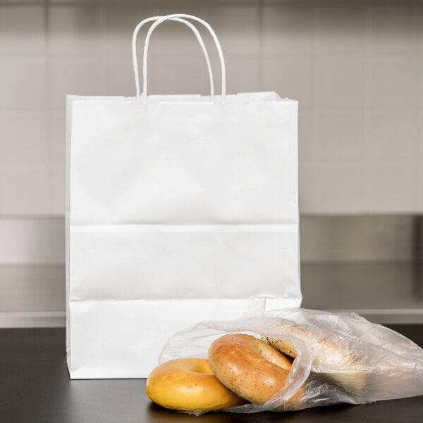 A white paper shopping bag with handles sitting next to plastic bags of bagels.
