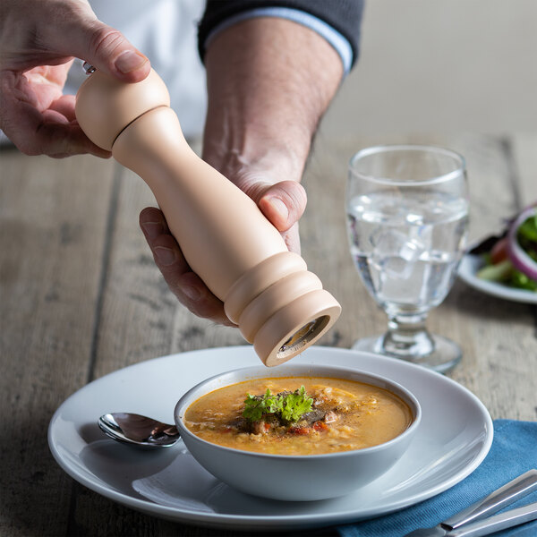 A person using an Acopa matte cream wooden pepper mill to pour pepper into a bowl of soup.