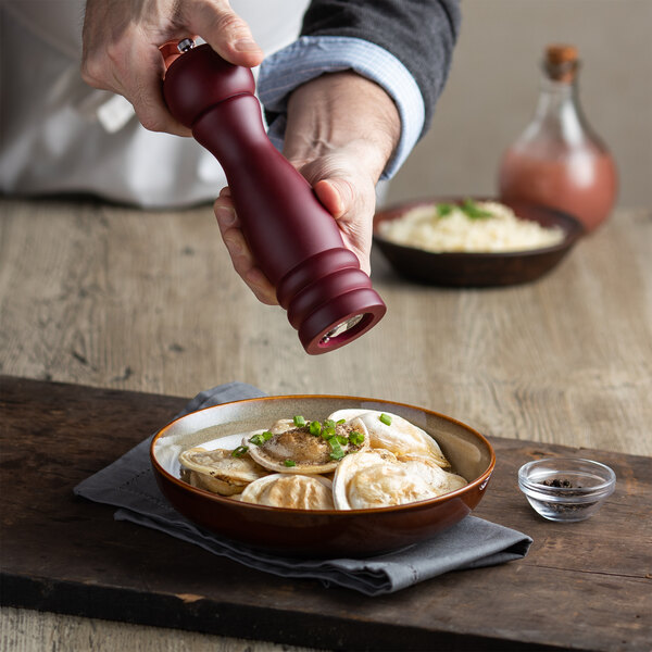 A person using an Acopa matte maroon wooden pepper mill to add pepper to a bowl of food.
