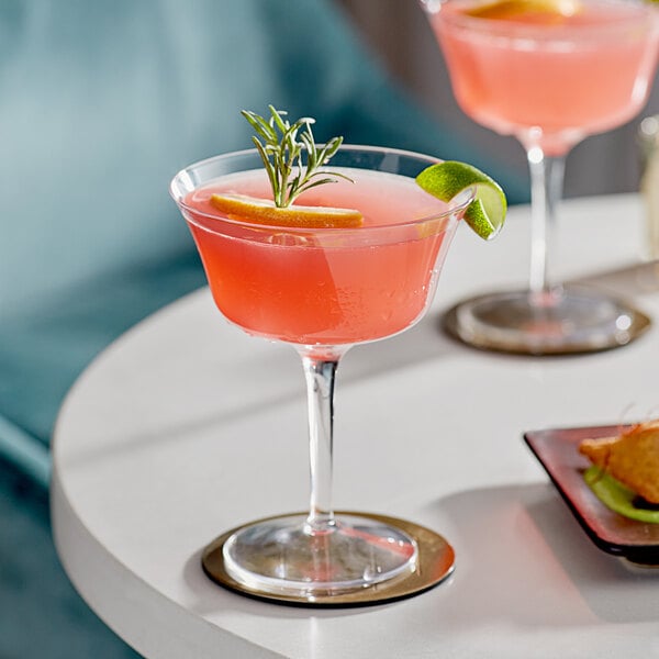 Two Acopa Deco flared coupe glasses filled with pink drinks, garnished with rosemary and lime, on a white table.