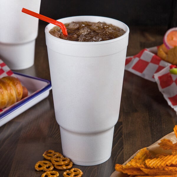 A white Dart foam cup with a straw in it on a table with a cup of soda and a bag of chips.