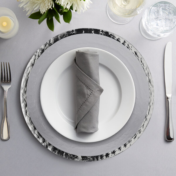A 10 Strawberry Street Alpine silver rim glass charger plate with a napkin and silverware on a table.