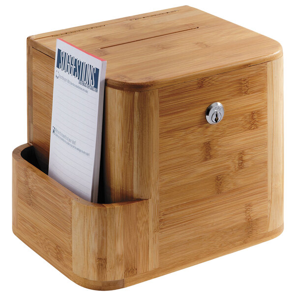 A Safco natural bamboo suggestion box on a counter with a piece of paper in it.