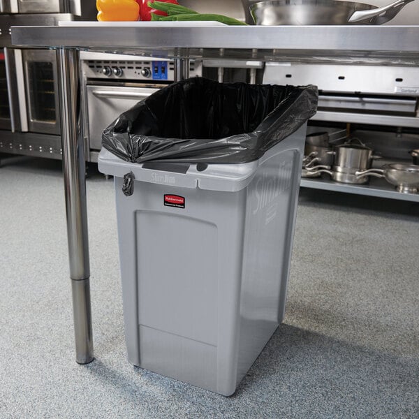 A grey Rubbermaid Slim Jim trash can under a black bag in a kitchen.