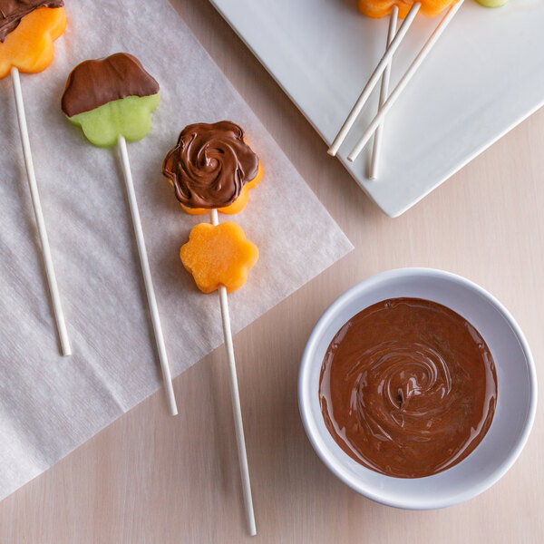 A plate of fruit skewers with chocolate on flower-shaped paper sticks.