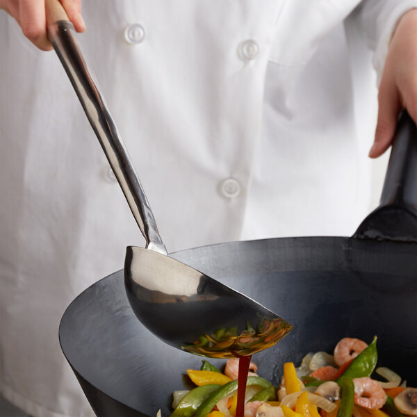 A person in a white coat using a Town medium wok ladle to pour sauce into a wok.