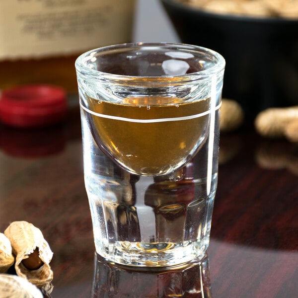A Libbey fluted shot glass with a .5 oz. pour line filled with a clear liquid next to a bowl of peanuts.