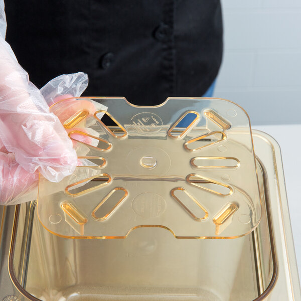A person in gloves using a Carlisle StorPlus amber high heat drain tray over a transparent container.
