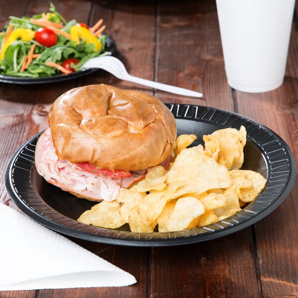 A Dart black foam plate with a sandwich, chips, and a salad on a table.