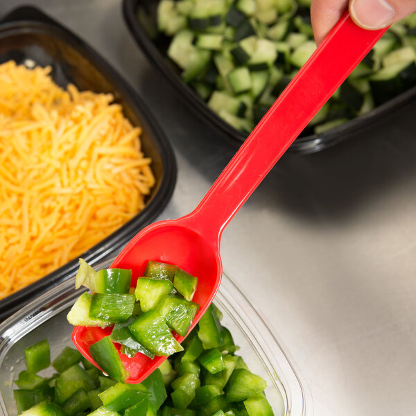A hand holding a Thunder Group red polycarbonate salad bar spoon over a bowl of chopped green peppers.