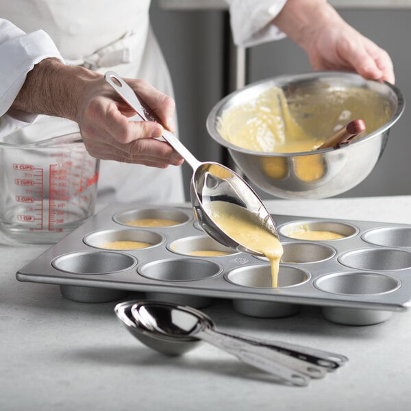 A person using Vollrath stainless steel measuring spoons to pour batter into a muffin tin.