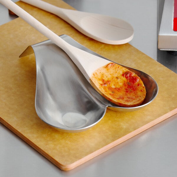 A Tablecraft brushed stainless steel double spoon rest holding a metal spoon and a wooden spoon with red sauce.