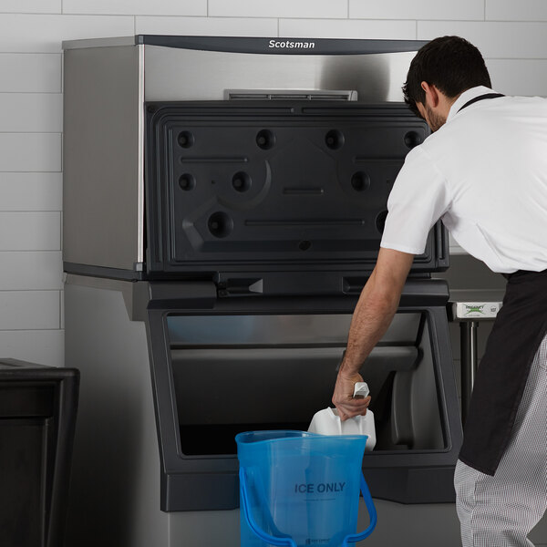 A man in a white shirt and apron using a blue plastic container to clean a Scotsman air cooled ice machine.