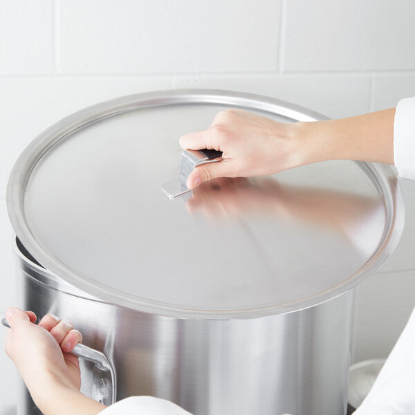 A person using a Vollrath domed aluminum pot cover with a metal handle on a large silver pot.