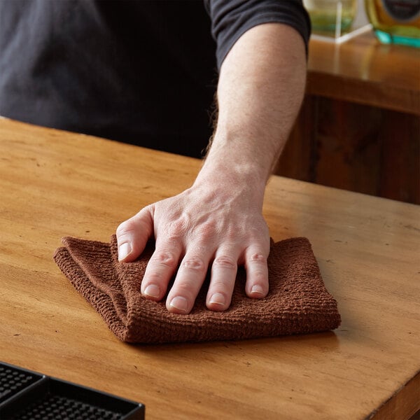 A hand using a brown Choice textured terry bar towel to wipe a table in a bar.